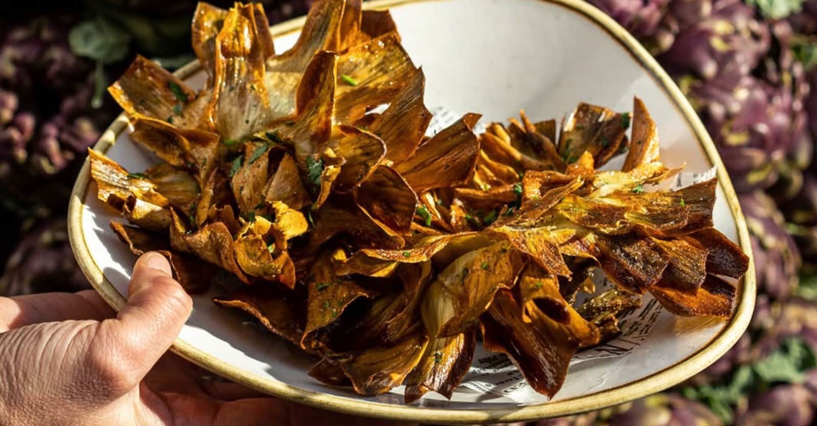 Foglie di carciofo fritte e croccanti impilate su un piatto bianco, cosparse di prezzemolo tritato.