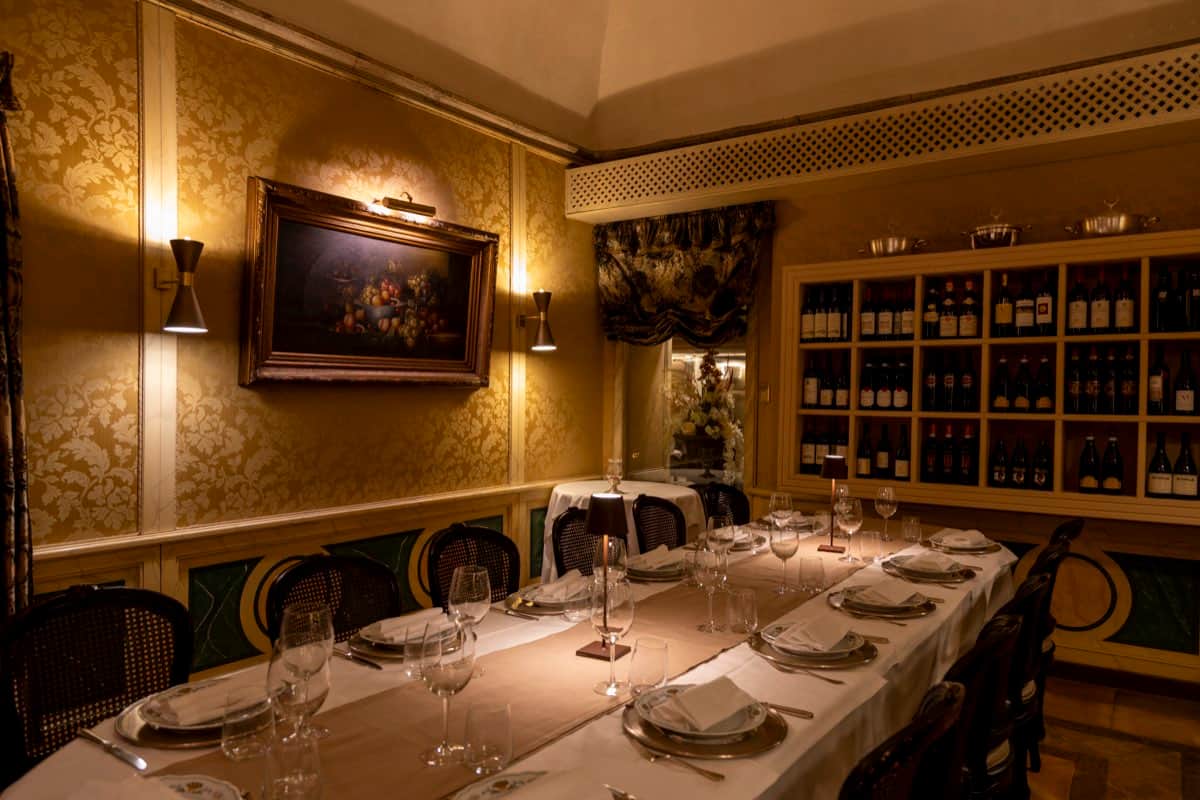 Sala da pranzo con tavolo lungo apparecchiato, pareti damascate dorate e libreria a vista con bottiglie di vino.