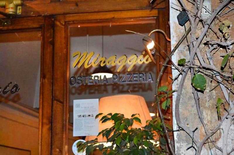 Vetrina con insegna dorata Miraggio e sotto la scritta Osteria Pizzeria, lampada accesa e piante verdi.