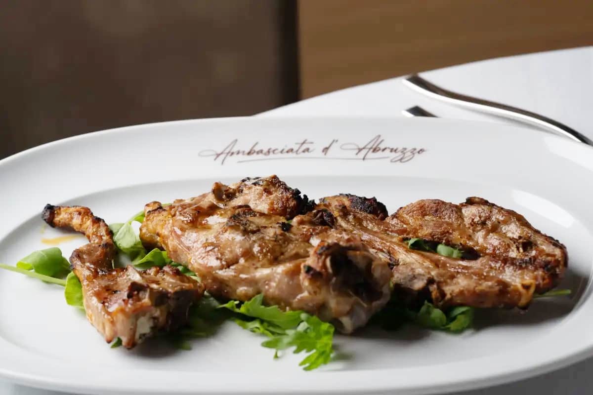 Costolette di carne grigliate su foglie verdi, in un piatto bianco con la scritta Ambasciata d'Abruzzo sul bordo.